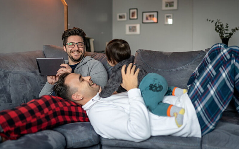 Ein homosexuelles Paar um die 30 liegt entspannt auf der Couch und spielt mit ihrem Baby.