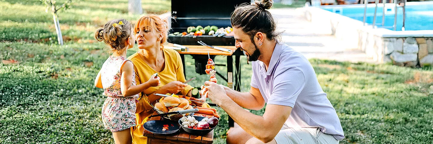 Gesund grillen: Ein Elternpaar bereitet mit seiner Tochter Gemüse, Brot und Würstchen für den Grill vor, der im Hintergrund steht.