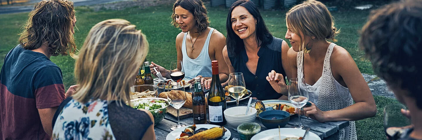 Eine Gruppe fröhlicher Personen sitzt an einem Gartentisch und genießt gesund Gegrilltes.