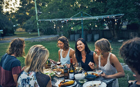 Eine Gruppe fröhlicher Personen sitzt an einem Gartentisch und genießt gesund Gegrilltes.