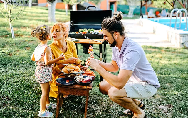 Gesund grillen: Ein Elternpaar bereitet mit seiner Tochter Gemüse, Brot und Würstchen für den Grill vor, der im Hintergrund steht.