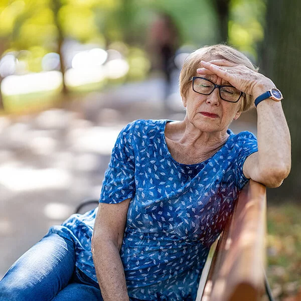 Eine Seniorin sitzt bei schönem Wetter auf einer Parkbank und greift sich mit der linken Hand an die Stirn.