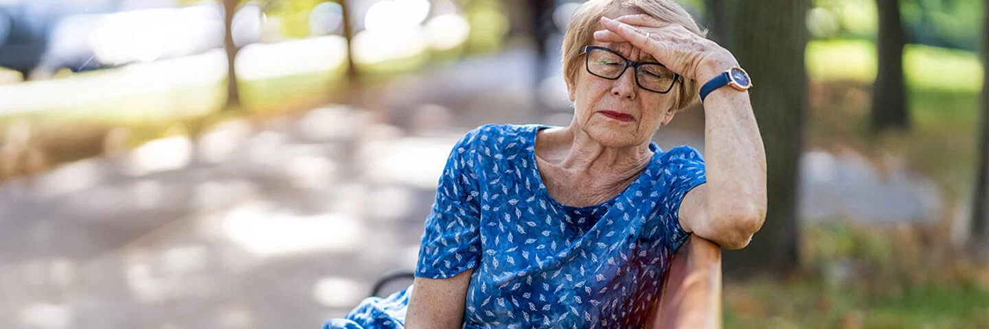 Eine Seniorin sitzt bei schönem Wetter auf einer Parkbank und greift sich mit der linken Hand an die Stirn.