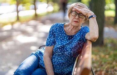 Eine Seniorin sitzt bei schönem Wetter auf einer Parkbank und greift sich mit der linken Hand an die Stirn.