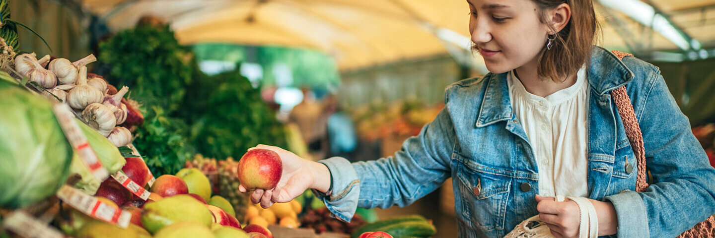 Junge Frau steht an einem Marktstand. Sie hält einen Apfel in der einen, ein gefülltes Einkaufsnetz in der anderen Hand.
