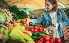 Junge Frau steht an einem Marktstand. Sie hält einen Apfel in der einen, ein gefülltes Einkaufsnetz in der anderen Hand.