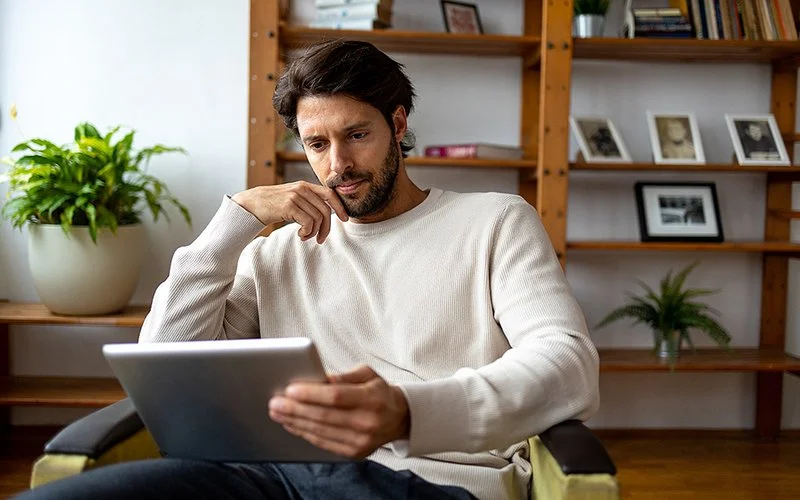 Ein Mann sitzt auf einem Sessel und guckt konzentriert auf ein Tablet.