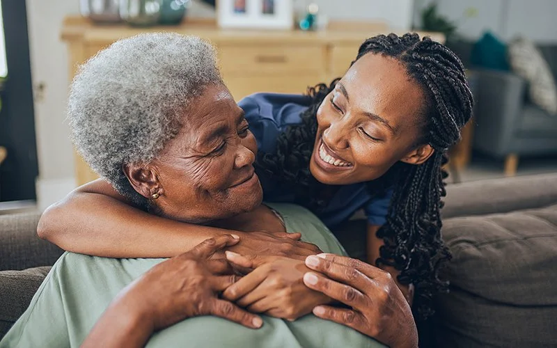 Eine Frau umarmt eine auf dem Sofa sitzende Person von hinten.