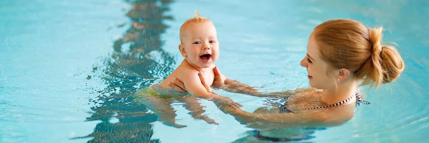 Eine blonde junge Frau schwimmt mit ihrem Baby in einem Schwimmbad und hält es an den Händen.