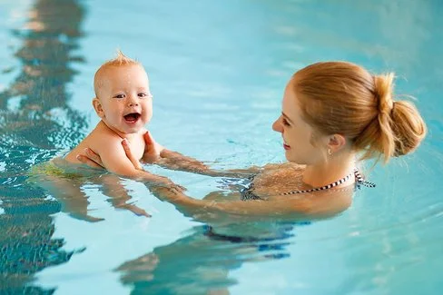 Eine blonde junge Frau schwimmt mit ihrem Baby in einem Schwimmbad und hält es an den Händen.