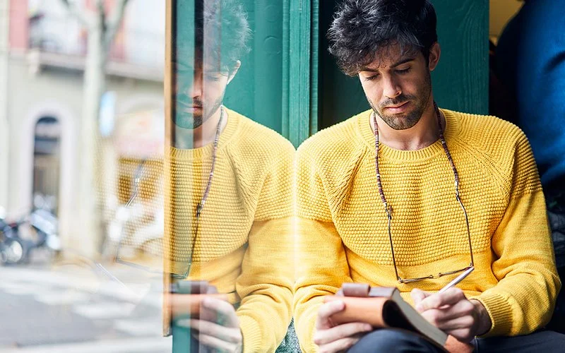 Ein jüngerer Mann mit einem gelben Pullover und dunklen Haaren sitzt am Fenster und notiert etwas in einem Notizbuch.