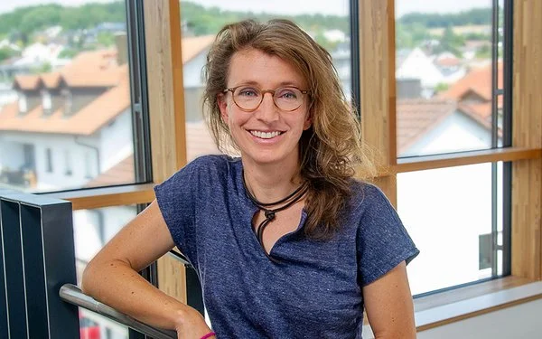 Dr. Barbara Eigenschenk steht vor einem großen Fenster, ihr Arm ruht auf einem Treppengeländer.