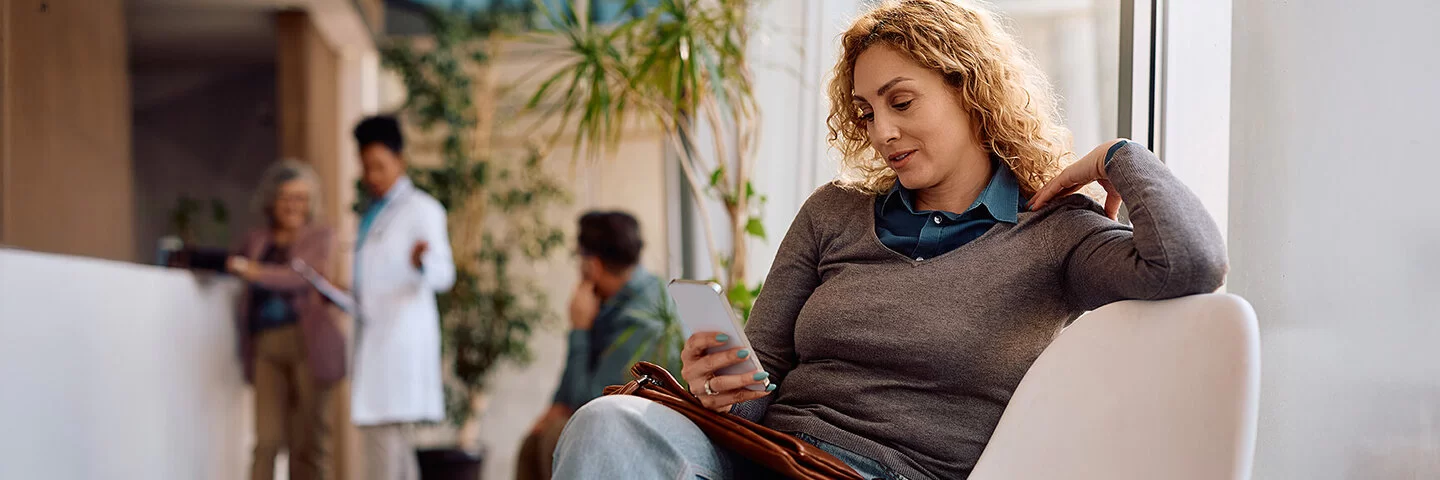 Eine Frau sitzt im Wartebereich einer Arztpraxis und schaut auf ihr Smartphone. Sie wirkt entspannt. Im Hintergrund sind weitere Personen zu sehen, darunter eine Ärztin im weißen Kittel, die mit anderen Patienten und Patientinnen spricht.