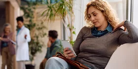 Eine Frau sitzt im Wartebereich einer Arztpraxis und schaut auf ihr Smartphone. Sie wirkt entspannt. Im Hintergrund sind weitere Personen zu sehen, darunter eine Ärztin im weißen Kittel, die mit anderen Patienten und Patientinnen spricht.