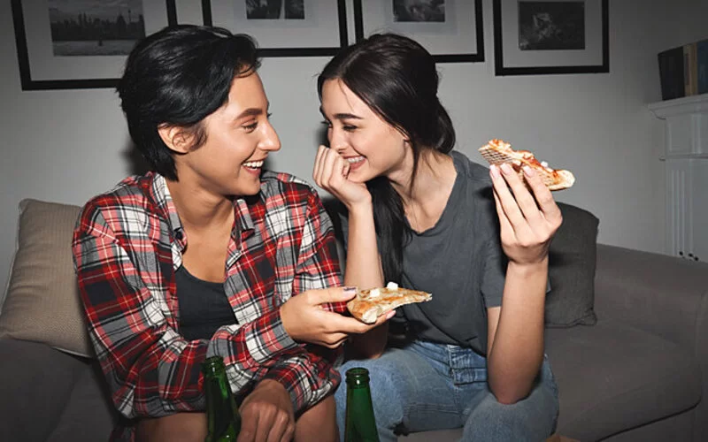 Zwei Freundinnen in einer Freundschaft Plus sitzen auf einem Sofa, lachen und essen Pizza.