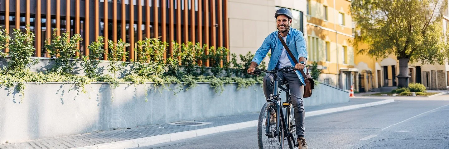 Ein Mann in Bürokleidung und mit Arbeitstasche fährt lächelnd mit dem Fahrrad durch die Stadt.