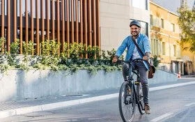 Ein Mann in Bürokleidung und mit Arbeitstasche fährt lächelnd mit dem Fahrrad durch die Stadt.