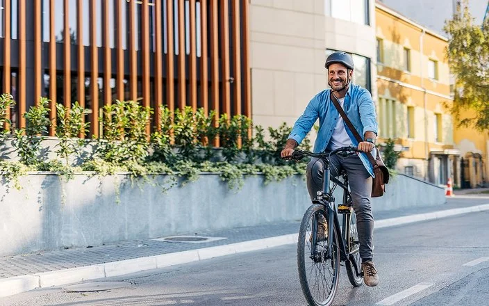 Ein Mann in Bürokleidung und mit Arbeitstasche fährt lächelnd mit dem Fahrrad durch die Stadt.