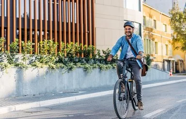 Ein Mann in Bürokleidung und mit Arbeitstasche fährt lächelnd mit dem Fahrrad durch die Stadt.