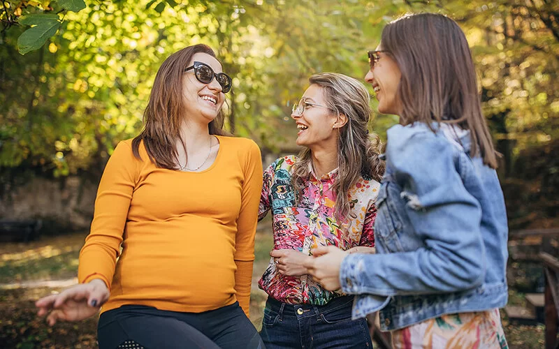 Drei junge Frauen, eine von ihnen schwanger, sitzen lachend und unterhaltend auf einer Parkbank bei angenehmen Temperaturen.