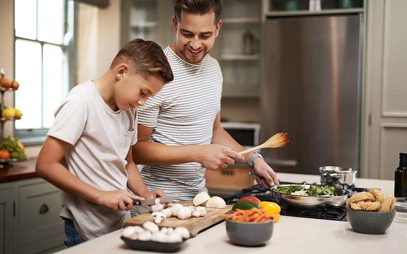 Ein Mann steht gemeinsam mit einem Jungen in der Küche und kocht auf einem Herd ein Gericht mit Champignons.