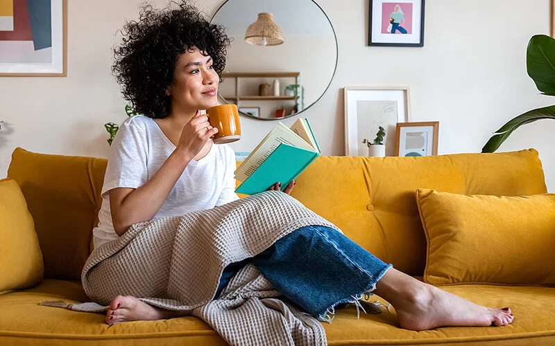 Eine junge Frau sitzt entspannt auf der Couch, hält ein Buch in der einen und einen Henkelbecher in der anderen Hand.