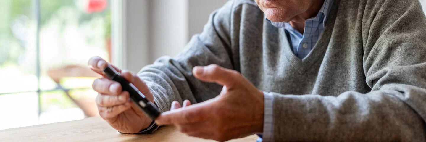 Ein Mann mit Brille sitzt an einem Holztisch und sticht sich mit einem Glucometer in den linken Zeigefinger.