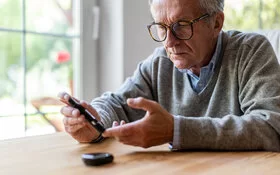 Ein Mann mit Brille sitzt an einem Holztisch und sticht sich mit einem Glucometer in den linken Zeigefinger.