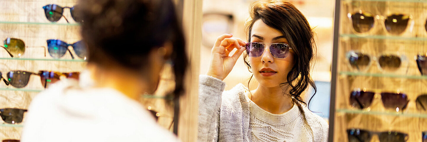 Eine Frau probiert im Brillengeschäft eine Sonnenbrille und schaut in einen Spiegel.