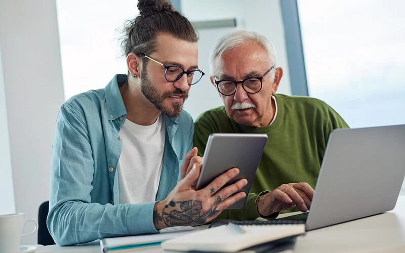 Jüngerer Mann erklärt einem älteren Mann am Laptop die Vorzüge der Telemedizin.