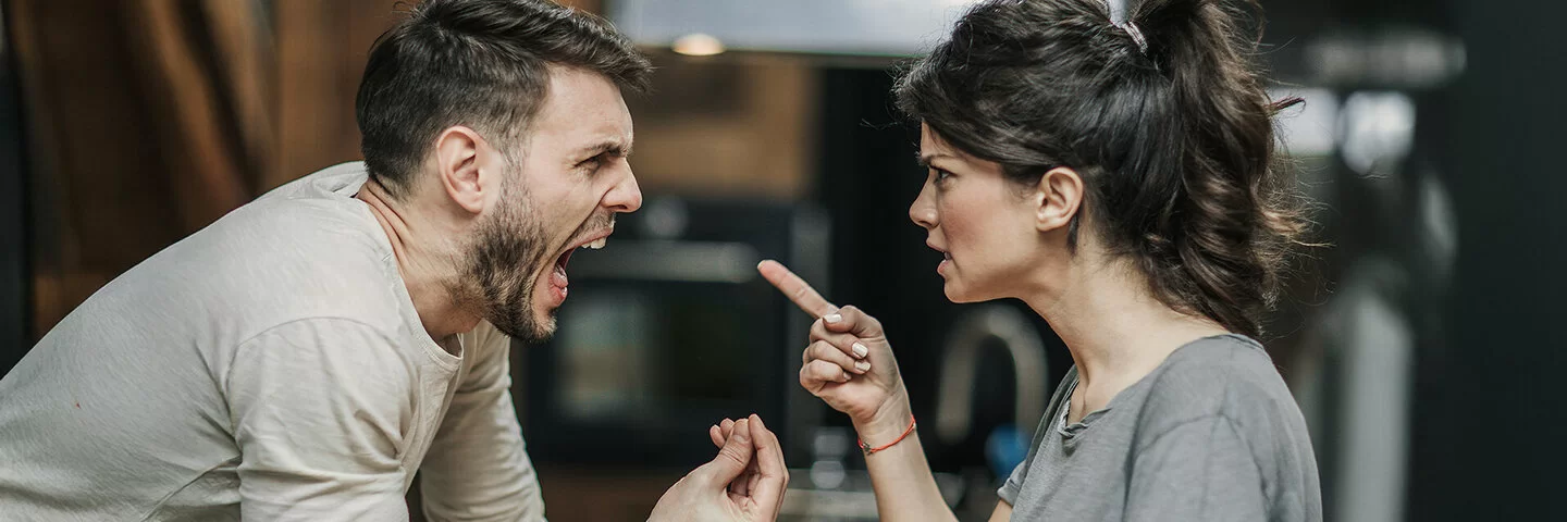 Ein Paar steht sich gegenüber. Beide streiten miteinander und sind sehr wütend. Die Frau richtet den Zeigefinger auf ihren Partner. Der Mann hat den Mund weit geöffnet und schreit. Mit den Fingern der rechten Hand macht er eine Drohgebärde.