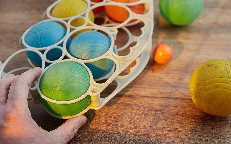 Bunte Boccia-Kugeln in einer Aufbewahrungsbox auf einem Holztisch, mit einer Hand, die eine Kugel herausnimmt.