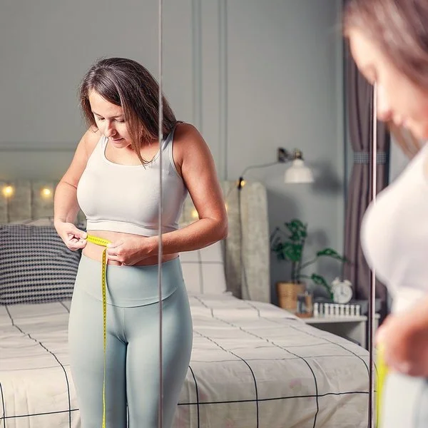 Eine junge Frau steht vor dem Spiegel und misst ihren Bauchumfang mit einem Maßband.