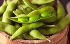 Frische grüne Edamame mit Schoten in einem Holzgefäß. Ein Schote ist offen, wodurch drei Edamamebohnen zu sehen sind.