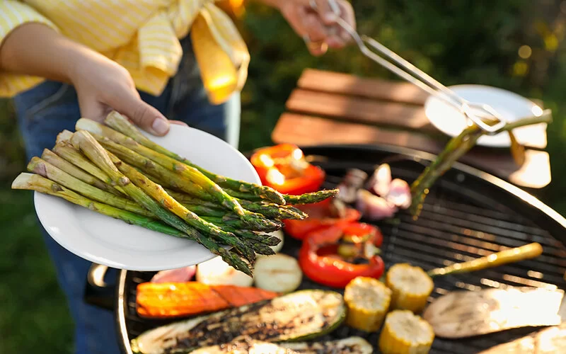 Eine Frau grillt im Freien Gemüse wie grünen Spargel, Mais, Paprika und Zucchini.