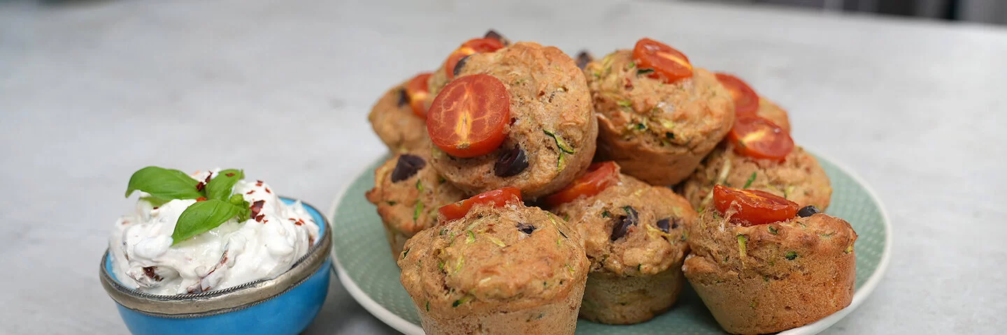 Gesunde Gemüsemuffins mit Käse nach einem Rezept von Felicitas Then, angerichtet auf einem hellgrünen Teller, garniert mit Tomaten. Daneben eine hellblaue Schale mit Dip, garniert mit einem Basilikumblatt.