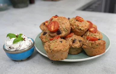 Gesunde Gemüsemuffins mit Käse nach einem Rezept von Felicitas Then, angerichtet auf einem hellgrünen Teller, garniert mit Tomaten. Daneben eine hellblaue Schale mit Dip, garniert mit einem Basilikumblatt.