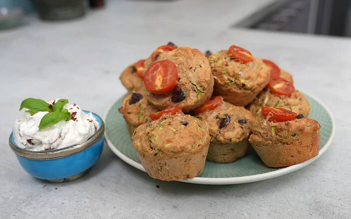 Gesunde Gemüsemuffins mit Käse nach einem Rezept von Felicitas Then, angerichtet auf einem hellgrünen Teller, garniert mit Tomaten. Daneben eine hellblaue Schale mit Dip, garniert mit einem Basilikumblatt.