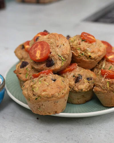Gesunde Gemüsemuffins mit Käse nach einem Rezept von Felicitas Then, angerichtet auf einem hellgrünen Teller, garniert mit Tomaten. Daneben eine hellblaue Schale mit Dip, garniert mit einem Basilikumblatt.