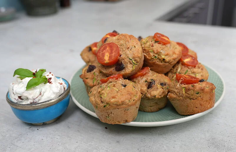 Gesunde Gemüsemuffins mit Käse nach einem Rezept von Felicitas Then, angerichtet auf einem hellgrünen Teller, garniert mit Tomaten. Daneben eine hellblaue Schale mit Dip, garniert mit einem Basilikumblatt.