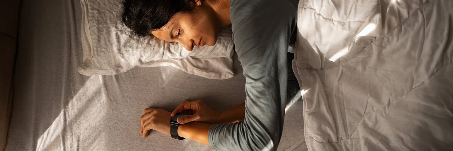 Eine Person liegt auf der Seite in einem Bett, der Oberkörper ist teilweise von einer hellen Decke bedeckt. Der rechte Arm liegt ausgestreckt auf der Matratze, das Handgelenk trägt eine Armbanduhr oder ein Fitnessarmband. Das Licht fällt schräg von links oben auf die Szene und erzeugt deutliche Schatten auf Bett und Decke. Die Haare der Person sind lang und dunkel.