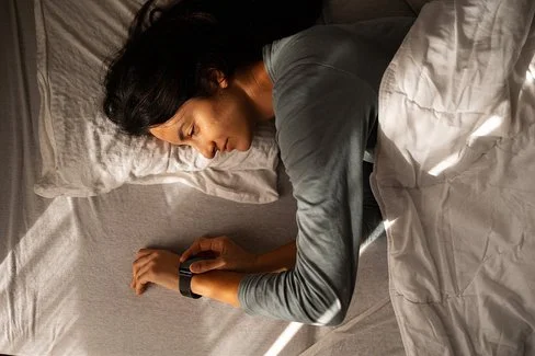 Eine Person liegt auf der Seite in einem Bett, der Oberkörper ist teilweise von einer hellen Decke bedeckt. Der rechte Arm liegt ausgestreckt auf der Matratze, das Handgelenk trägt eine Armbanduhr oder ein Fitnessarmband. Das Licht fällt schräg von links oben auf die Szene und erzeugt deutliche Schatten auf Bett und Decke. Die Haare der Person sind lang und dunkel.