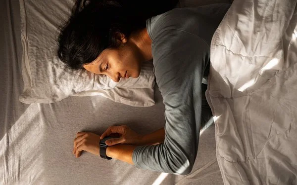 Eine Person liegt auf der Seite in einem Bett, der Oberkörper ist teilweise von einer hellen Decke bedeckt. Der rechte Arm liegt ausgestreckt auf der Matratze, das Handgelenk trägt eine Armbanduhr oder ein Fitnessarmband. Das Licht fällt schräg von links oben auf die Szene und erzeugt deutliche Schatten auf Bett und Decke. Die Haare der Person sind lang und dunkel.
