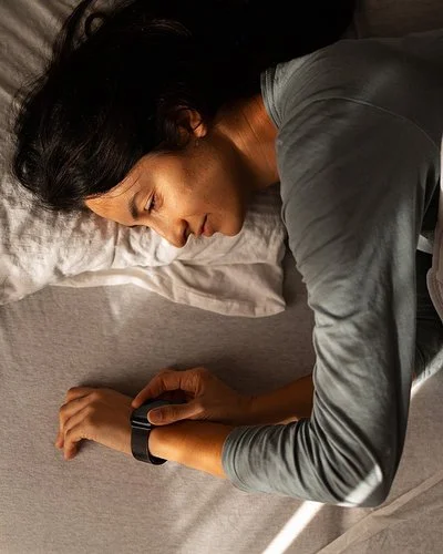 Eine Person liegt auf der Seite in einem Bett, der Oberkörper ist teilweise von einer hellen Decke bedeckt. Der rechte Arm liegt ausgestreckt auf der Matratze, das Handgelenk trägt eine Armbanduhr oder ein Fitnessarmband. Das Licht fällt schräg von links oben auf die Szene und erzeugt deutliche Schatten auf Bett und Decke. Die Haare der Person sind lang und dunkel.