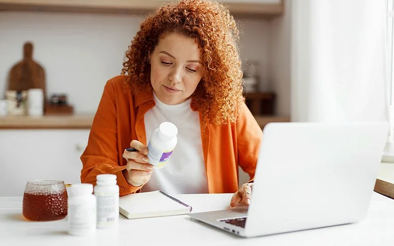 Eine junge Frau sitzt vor ihrem Rechner. In der rechten Hand hält sie eine Plastikdose mit einem Nahrungsergänzungsmittel und liest aufmerksam die Hinweise auf dem Etikett.