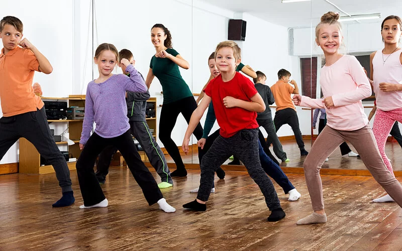 Eine Gruppe von Mädchen und Jungen führt unter Anleitung einer Trainerin in einem Tanzstudio choreografierte Tanzschritte aus.