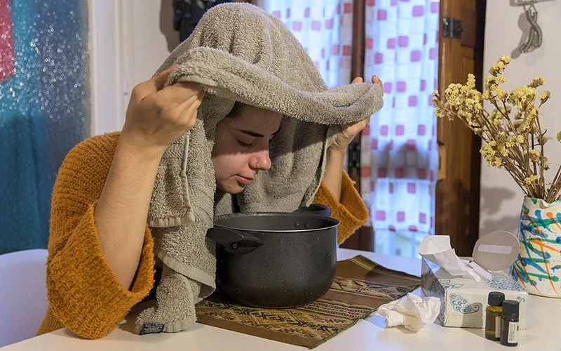 Eine Frau hat ein Handtuch über ihren Kopf gelegt und beugt ihn über einen Kochtopf, um den Dampf zu inhalieren.