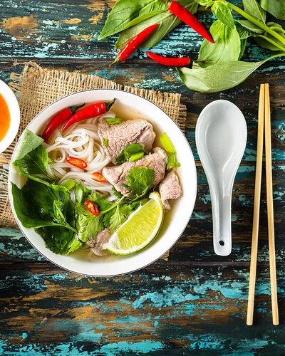 Eine vietnamesische Suppe steht auf einem hölzernen Untergrund, links daneben stehen eine Schüssel mit frischen Zuaten und eine Soße.