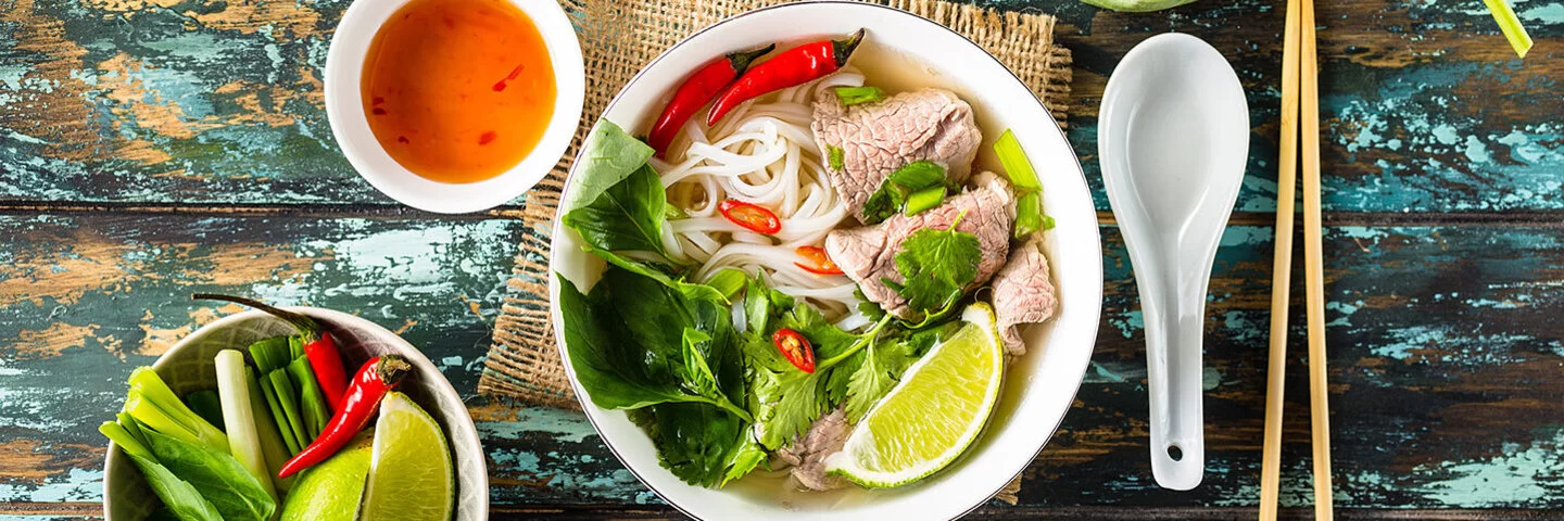 Eine vietnamesische Suppe steht auf einem hölzernen Untergrund, links daneben stehen eine Schüssel mit frischen Zuaten und eine Soße.