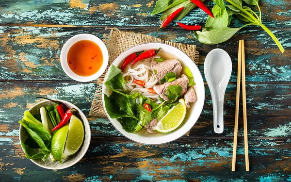Eine vietnamesische Suppe steht auf einem hölzernen Untergrund, links daneben stehen eine Schüssel mit frischen Zuaten und eine Soße.